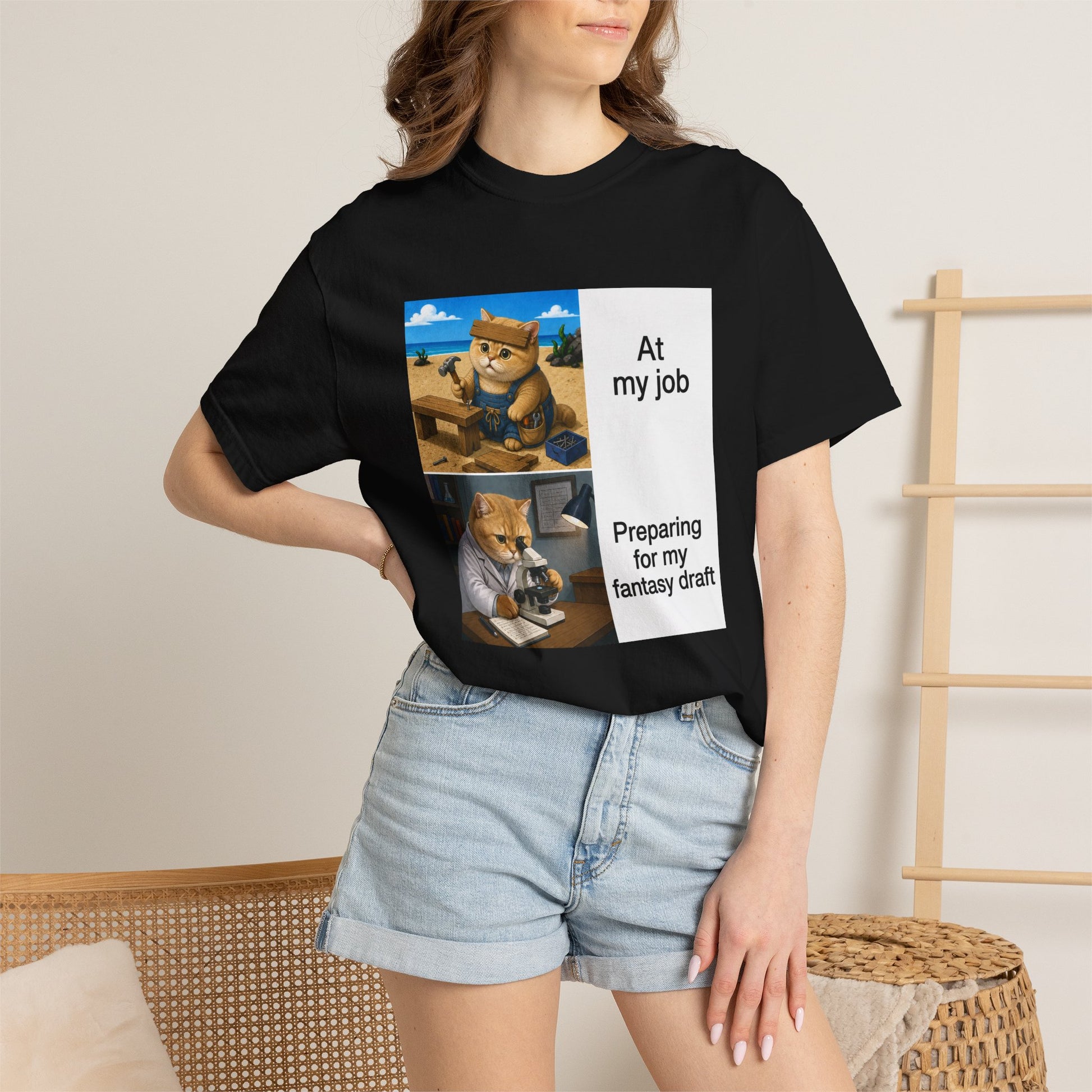 Black Comfort Colors 1717 t-shirt with a square two-panel print on an off-white card across the chest: an orange cat in denim overalls hammering a nail into a wooden bench on a beach, captioned At my job, above the same cat in a white lab coat hunched over a microscope beside a notebook of stats, captioned Preparing for my fantasy draft.