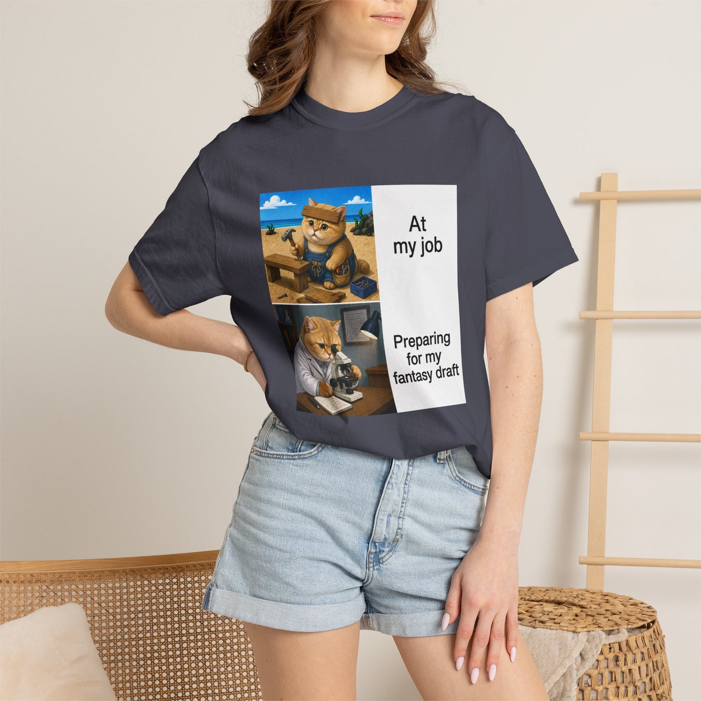 Navy Comfort Colors 1717 t-shirt with a square two-panel print on an off-white card across the chest: an orange cat in denim overalls hammering a nail into a wooden bench on a beach, captioned At my job, above the same cat in a white lab coat hunched over a microscope beside a notebook of stats, captioned Preparing for my fantasy draft.
