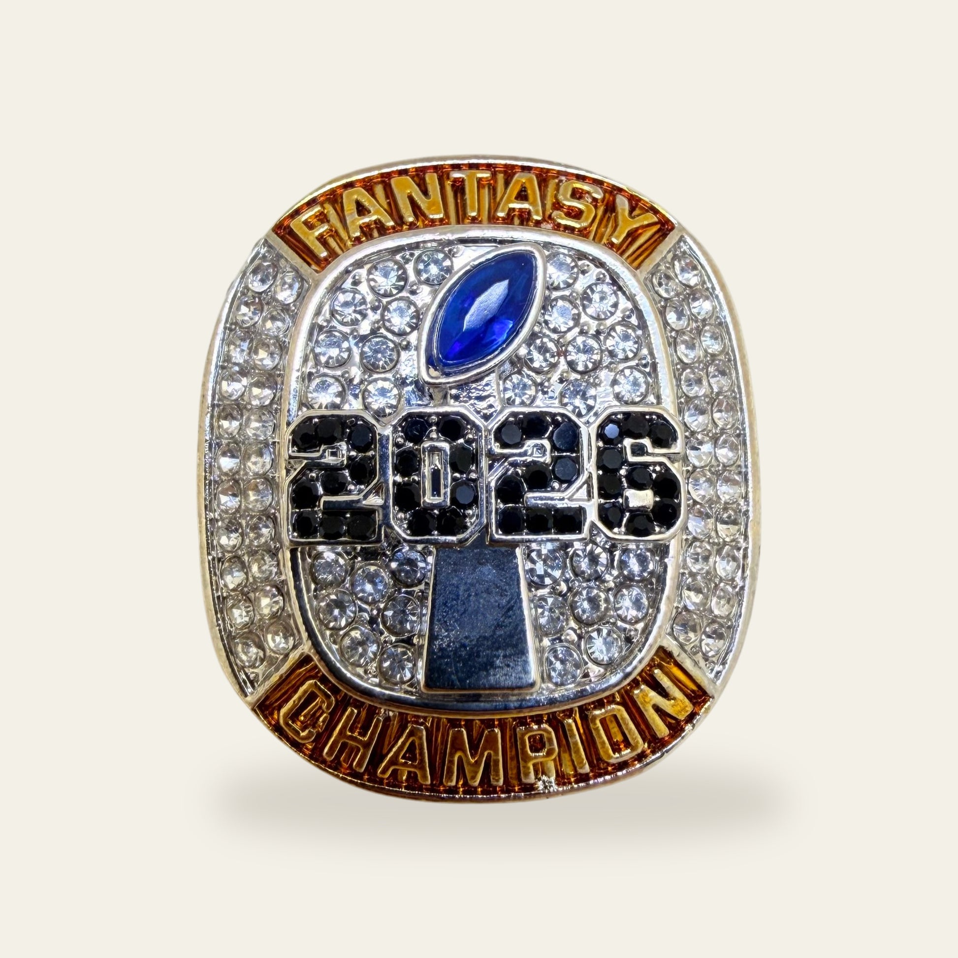 Fantasy football championship ring, face view, with gold FANTASY and CHAMPION banners around a clear stone paved face, a blue marquise football stone at the top, and 2026 in black stones over a silver trophy.