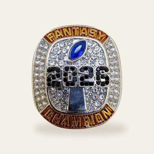 Fantasy football championship ring, face view, with gold FANTASY and CHAMPION banners around a clear stone paved face, a blue marquise football stone at the top, and 2026 in black stones over a silver trophy.