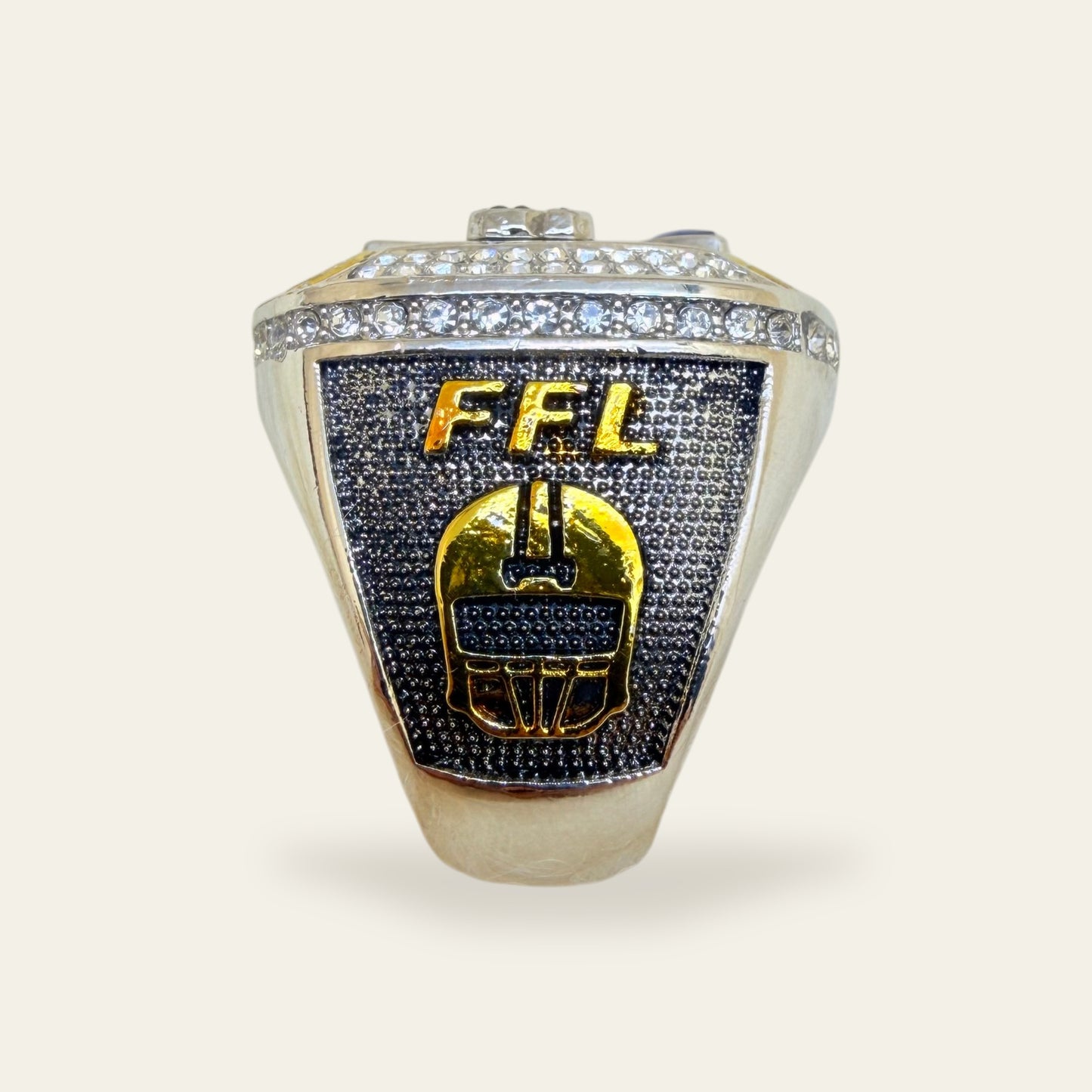Fantasy football championship ring, left shoulder view, with gold FFL letters above a gold football helmet on a dark textured panel, under a row of clear stones.