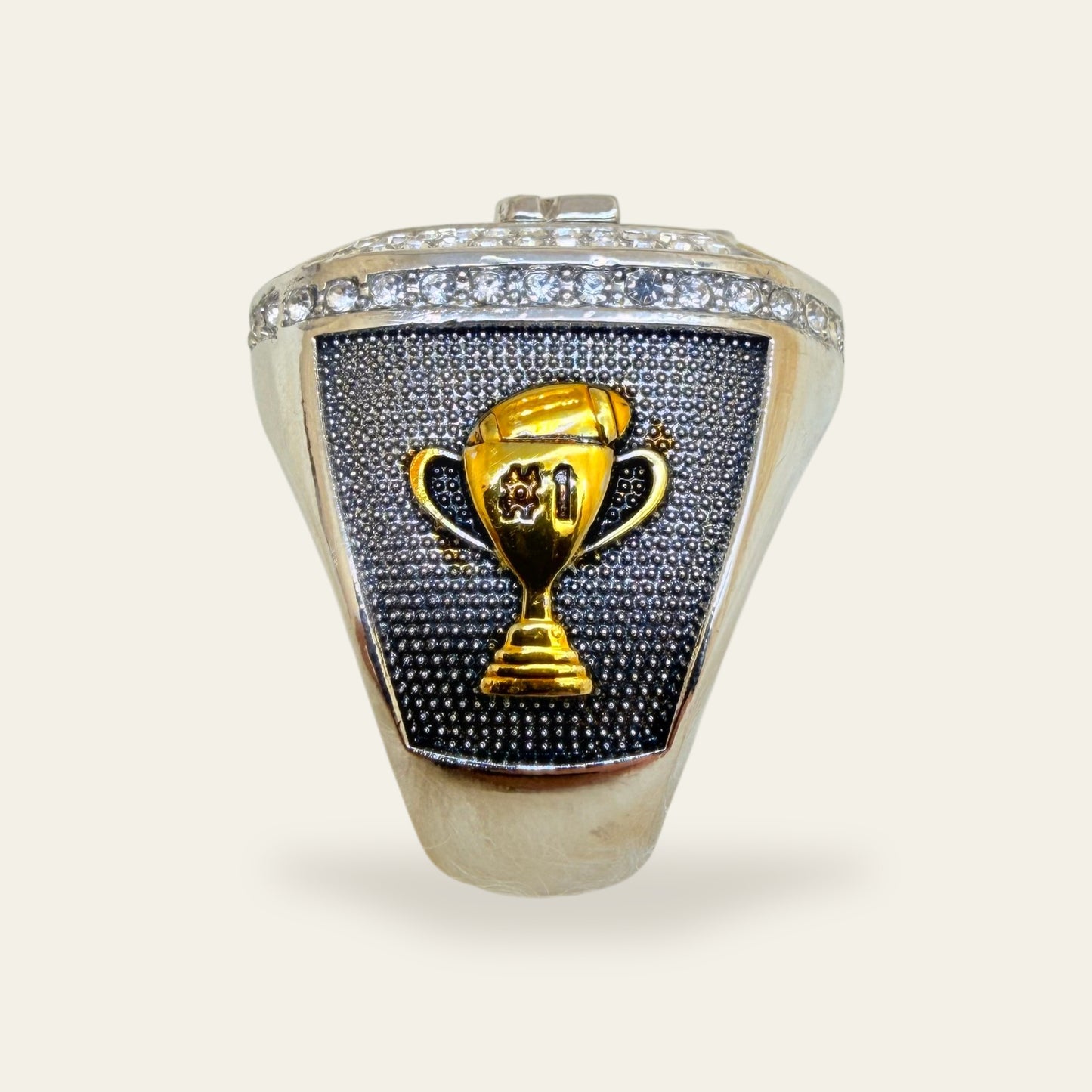 Fantasy football championship ring, right shoulder view, with a gold number one trophy topped by a football on a dark textured panel, under a row of clear stones.