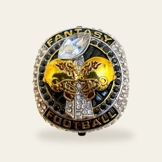 Black and white fantasy football championship ring, face view, with gold FANTASY and FOOTBALL banners, two gold helmets meeting over a clear stone trophy, a large clear marquise football stone at the top, and rows of black and clear stones around the edge.
