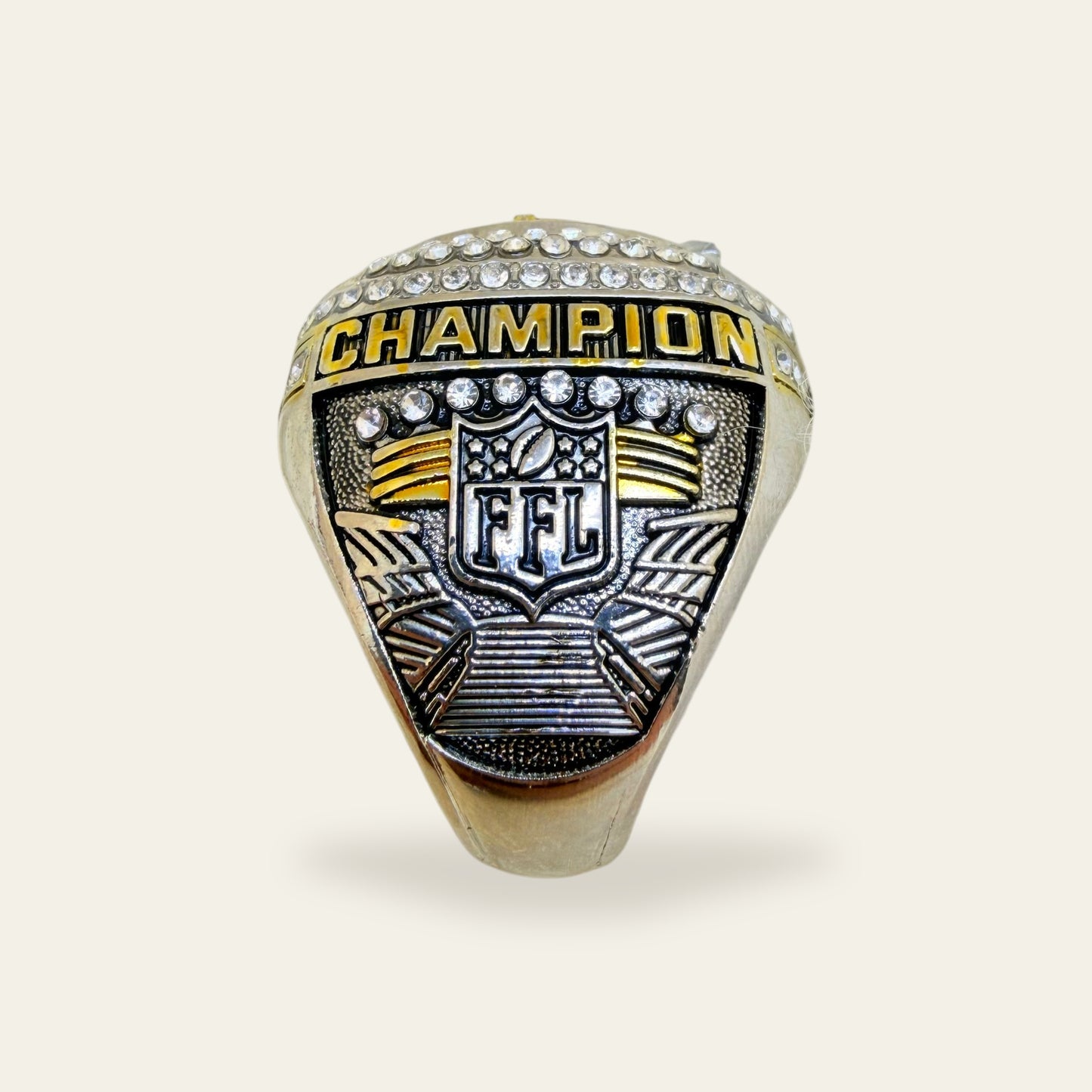 Black and white fantasy football championship ring, right shoulder view, with a gold CHAMPION rail, an FFL shield crest above stadium steps, and an arc of clear stones on a black pebbled panel.