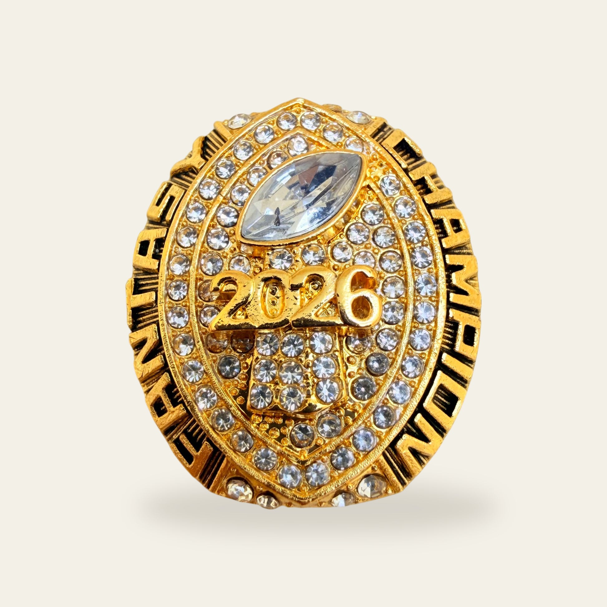 All gold fantasy football championship ring, face view, with a football shaped face paved in clear stones, a clear marquise football stone at the top, raised 2026 across the middle, a stone set trophy below, and FANTASY and CHAMPION along the sides.