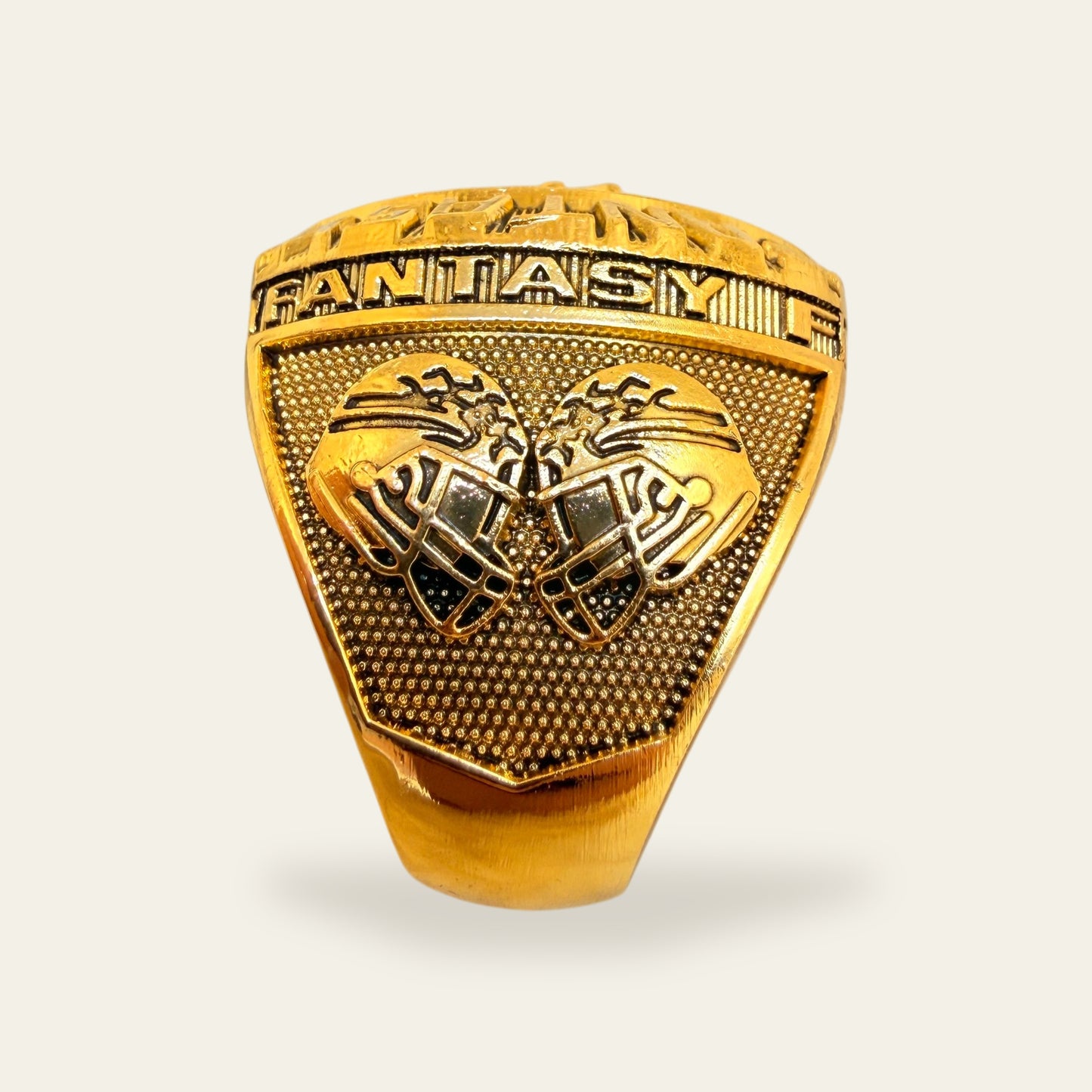 All gold fantasy football championship ring, left shoulder view, with two football helmets meeting on a stippled gold panel under a FANTASY rail.