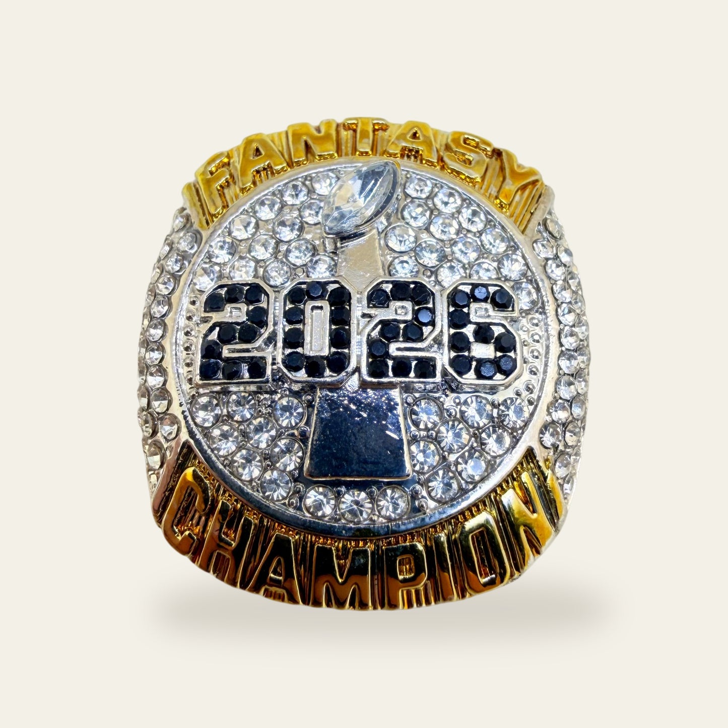 Two tone fantasy football championship ring, face view, with gold FANTASY and CHAMPION lettering around a clear stone paved silver face, a marquise football stone at the top, and 2026 in black stones over a silver trophy.