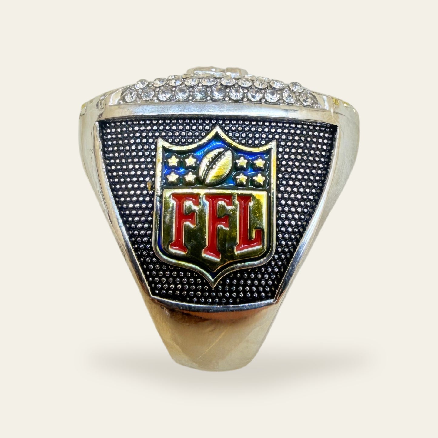 Two tone fantasy football championship ring, right shoulder view, with a red, blue, and gold FFL shield crest on a black stippled panel, under a row of clear stones.