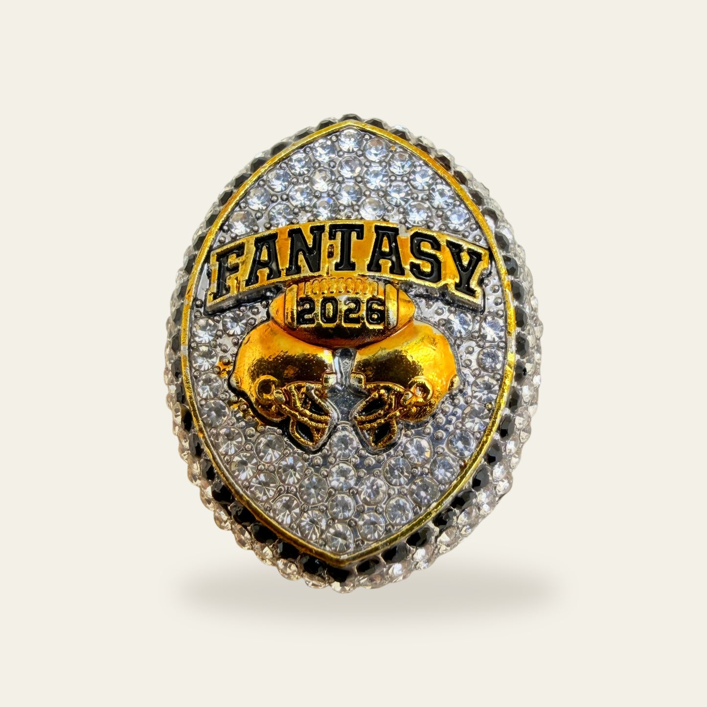 Whiteout fantasy football championship ring, face view, with a football shaped face paved in clear stones inside a gold frame, a gold trimmed FANTASY banner, a gold football carrying 2026, and two gold helmets meeting at the center.