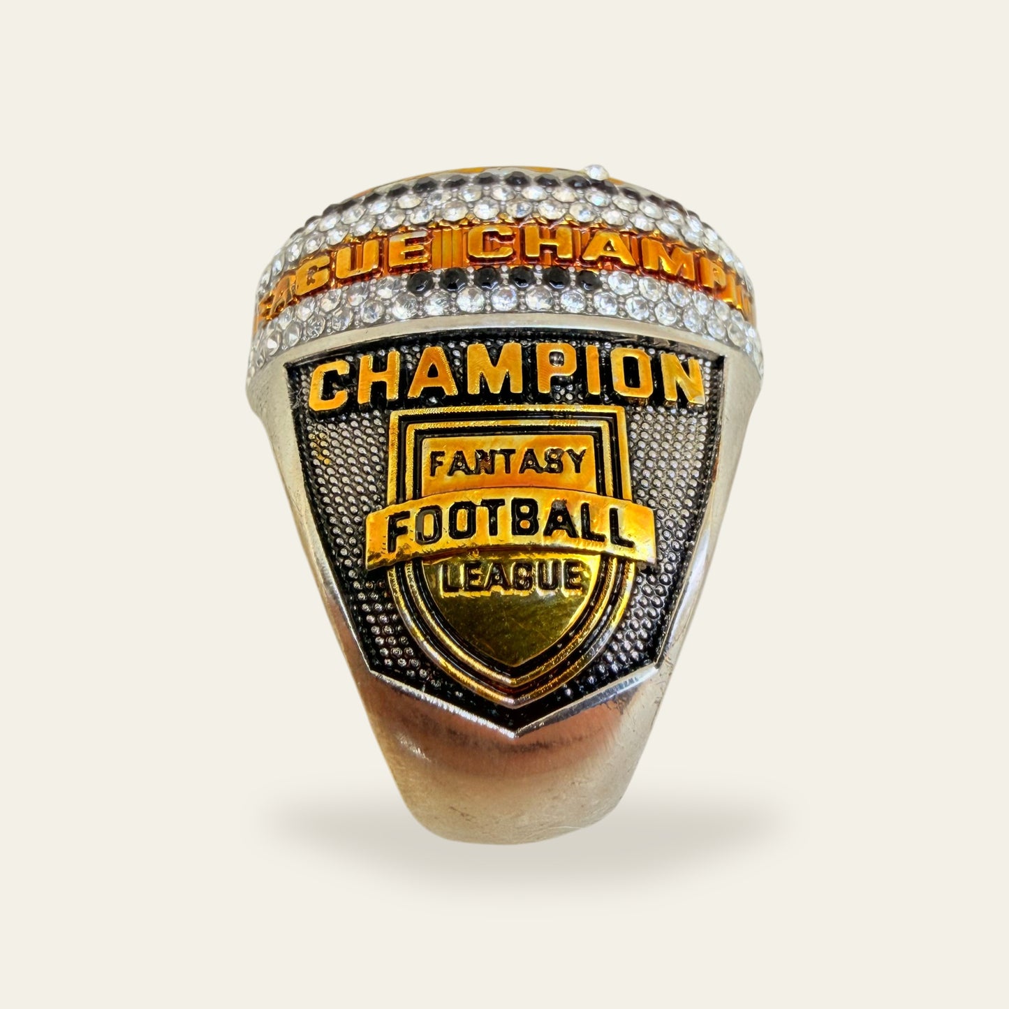 Whiteout fantasy football championship ring, left shoulder view, with gold CHAMPION letters above a Fantasy Football League shield crest, under a lettered rail set between rows of clear stones.