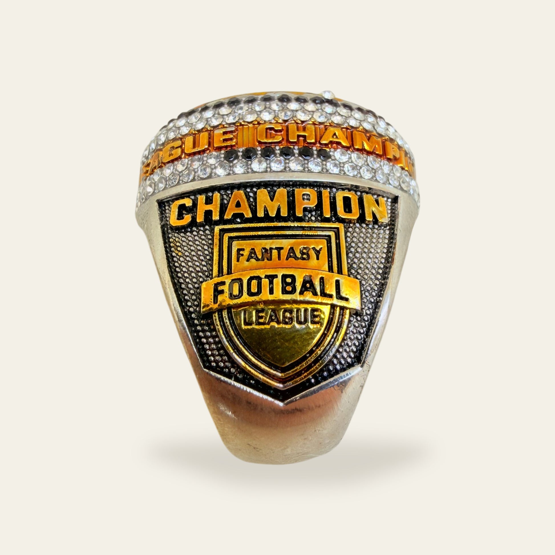 Whiteout fantasy football championship ring, left shoulder view, with gold CHAMPION letters above a Fantasy Football League shield crest, under a lettered rail set between rows of clear stones.