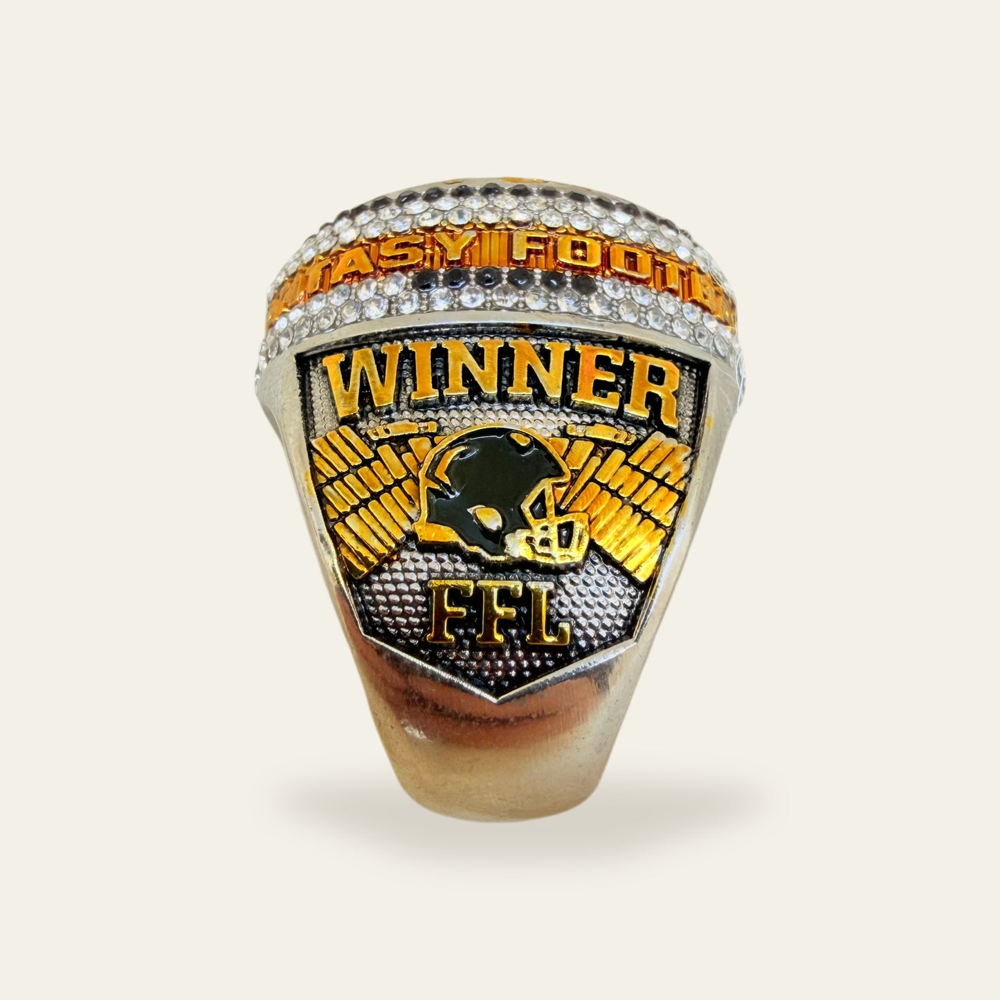 Whiteout fantasy football championship ring, right shoulder view, with gold WINNER letters above a black helmet framed by gold wings and FFL below, under a lettered rail set between rows of clear stones.